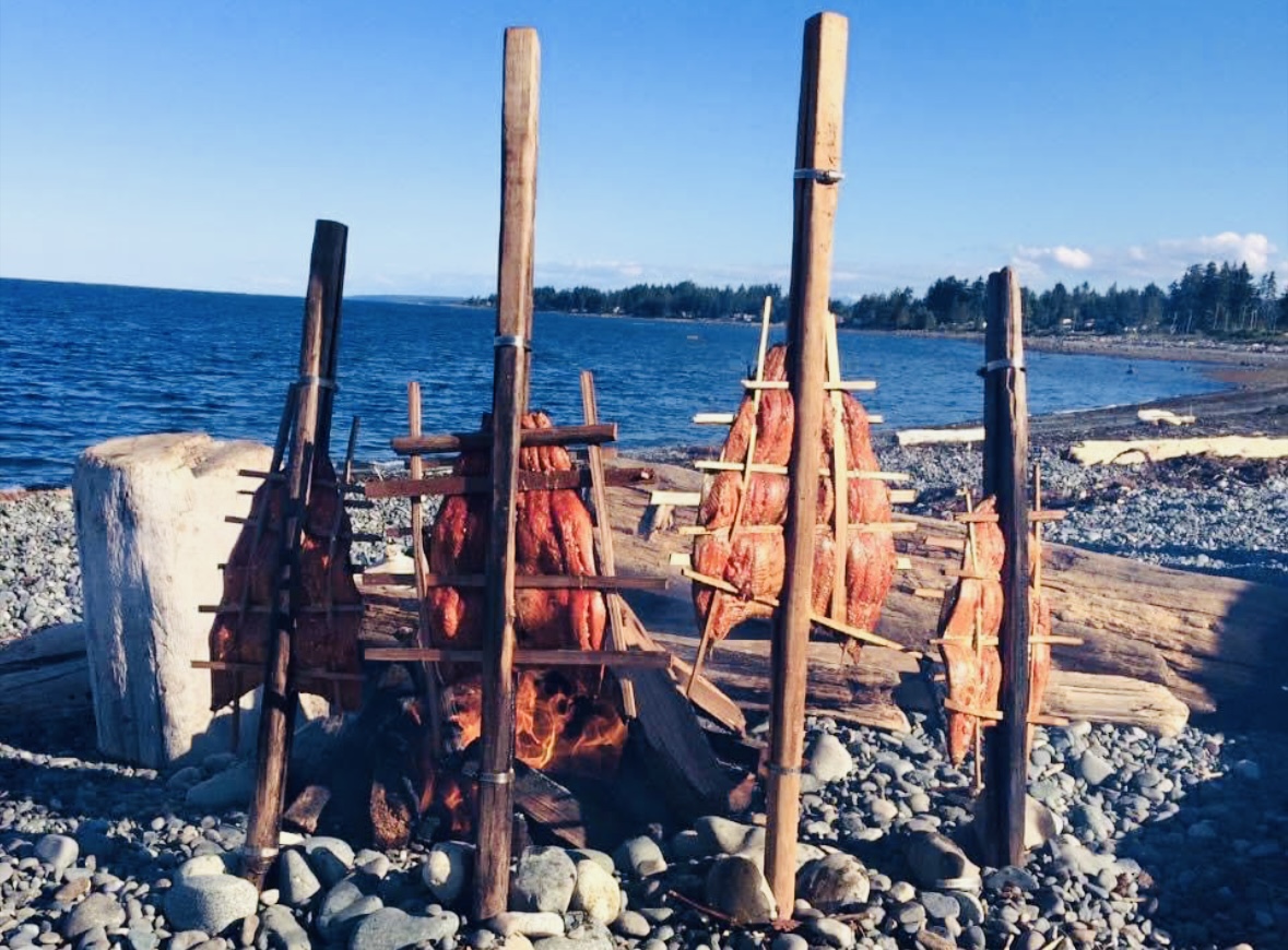 Four sets of sticks holding fish, stuck into the ground roasting next to a campfire. The campfire is on a rocky beach with a view of the surrounding forest and ocean.